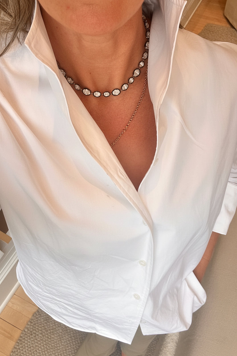 A close-up of a person wearing a white button-down shirt and layered necklaces, including a silver-toned beaded choker and a delicate gold chain.
