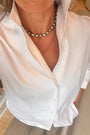 A close-up of a person wearing a white button-down shirt and layered necklaces, including a silver-toned beaded choker and a delicate gold chain.